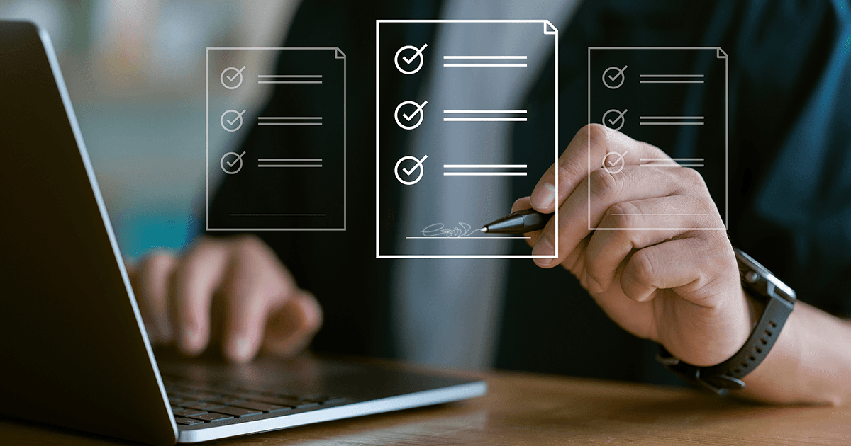 Hand holding pen checking off items on digital patent licensing checklist documents with laptop on wooden desk