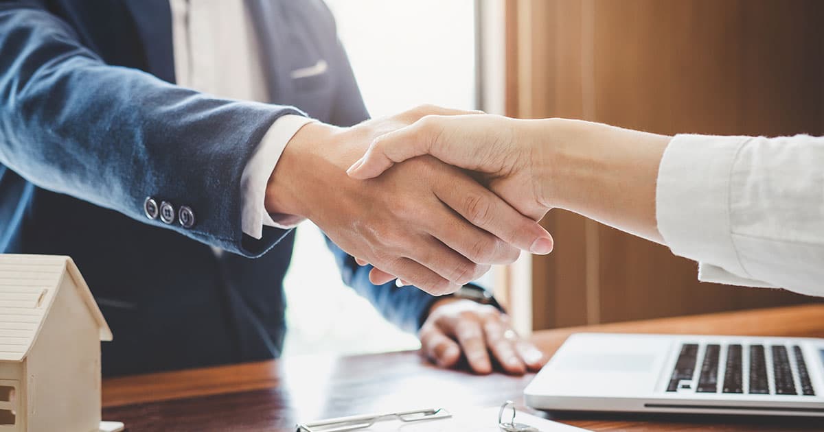 Two business professionals shaking hands over a desk with laptop and wooden house model, finalizing a patent sale agreement